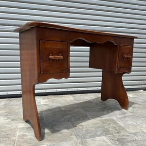 May include: A vintage wooden desk with a warm brown finish. The desk features two drawers with wooden handles and a scalloped edge along the top. The legs have a unique curved design. A classic piece of furniture.