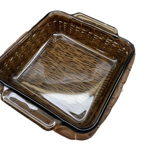May include: A square, clear glass baking dish with a brown woven wicker basket. The dish has a rectangular shape with rounded corners and handles. The basket is made of woven brown material.