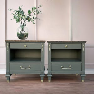 May include: Two matching nightstands in a classic style. The nightstands are painted a muted green with a dark wood top. Each has a drawer with a decorative handle and an open shelf. The legs have gold accents, and the drawer has a gold flower-shaped knob.