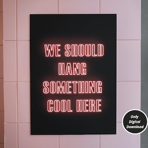 Op de afbeelding: Een zwarte poster met de tekst "WE SHOULD HANG SOMETHING COOL HERE" in roze neonletters. De poster hangt aan een roze betegelde muur. De tekst "Only Digital Download" staat in witte letters op een zwarte cirkel in de rechterbenedenhoek van de poster.