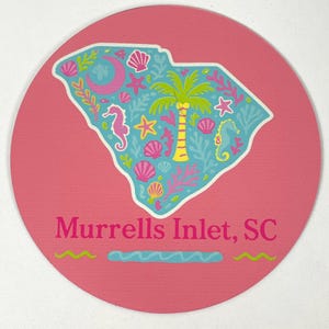 May include: A round, pink coaster featuring a teal outline of South Carolina filled with seashell, seahorse, and palm tree illustrations. The text "Murrells Inlet, SC" is printed in pink.