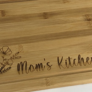 May include: A natural bamboo cutting board with a floral design and the text "Mom's Kitchen" engraved on the surface.