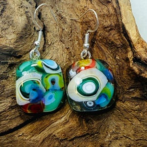 May include: A pair of square, dichroic glass earrings with sterling silver ear wires. The colorful glass features swirls of blue, green, white, red, and yellow. The earrings are displayed on a piece of textured, brown wood.