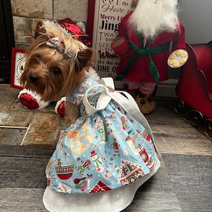 May include: A small, brown and white Yorkshire Terrier dog wearing a blue and white Christmas-themed dress with a red sequined Santa hat. The dog is sitting in front of a Santa Claus figurine.