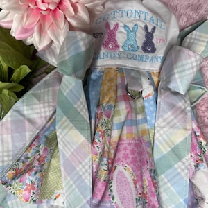 May include: A white dress with a pastel plaid pattern and floral print. The dress has a white bodice with the text "COTTONTAIL EST. 1779 CANDY COMPANY" embroidered on it. Three embroidered bunnies are in the text. The dress is tied with a blue ribbon.
