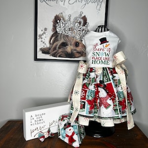 May include: A white dress with a snowman graphic and the text "There is SNOW PLACE LIKE HOME". The dress has a red and green plaid skirt with a white snowflake pattern. The dress is on a mannequin and there is a sign that says "A house is not a home without paw prints".