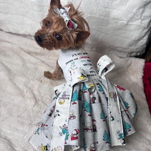 May include: A small dog wearing a festive Christmas dress and matching bow. The dress is gray with a pattern of cartoon characters, Christmas trees, and snow. The shirt has the text "IT'S NOT WHAT UNDER THE CHRISTMAS TREE THAT MATTERS IT'S WHO'S ROUND IT."