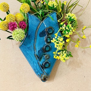 May include: A blue ceramic wall vase with a unique design and five round accents. The vase is filled with yellow and green artificial flowers.