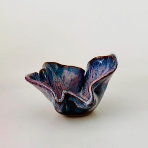 May include: A small ceramic bowl with a wavy, organic rim. The glaze features a blend of deep blue, purple, and soft pink tones with a glossy finish. The bowl has a flared, petal-like shape that curves upward, creating a distinct, irregular silhouette.