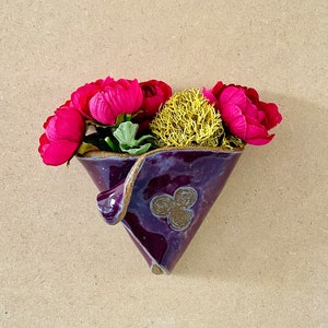 May include: A purple ceramic wall pocket vase with a gold rim, holding vibrant pink flowers and a yellow accent. The vase has a triangular shape and a decorative swirl design. The background is a light brown.