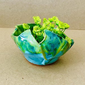 May include: A handmade ceramic bowl with a unique, folded design. The bowl features a blend of turquoise, green, and yellow glaze, creating a marbled effect. It is filled with bright green, leafy sprigs, adding a natural touch to the decorative piece.