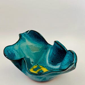 May include: A handcrafted ceramic bowl with a unique, organic shape. The bowl features a glossy teal glaze with darker blue accents and a yellow pattern at the base. The rim has a wavy design, adding to its artistic appeal.