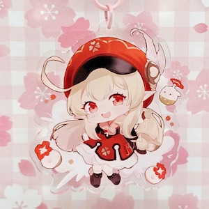 May include: A pink and white anime character keychain with a red hat and a white dress. The character is holding a small white bird in its hand. The keychain is surrounded by pink cherry blossoms.