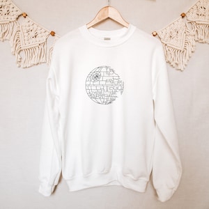 May include: White sweatshirt featuring a black graphic of the Death Star. The Death Star design is a detailed, geometric illustration. The sweatshirt is hanging on a wooden hanger against a neutral background.
