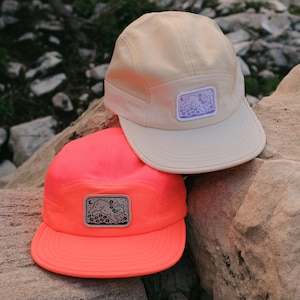 May include: Two five-panel caps, one in cream and one in neon orange, each featuring a rectangular patch on the front. The patches display a line-art illustration of a small animal running through flowers under a crescent moon and stars.