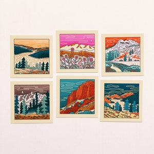 May include: Set of six illustrated cards featuring different landscape scenes. Each card has a colorful outline drawing of a mountain range, trees, and a body of water. The cards are printed on a cream-colored background.