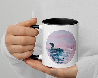 May include: White ceramic mug with a black rim and handle. The mug features a watercolour illustration of a loon swimming in a lake with a pink and purple sunset in the background. The text "be still" is written in cursive on the mug.