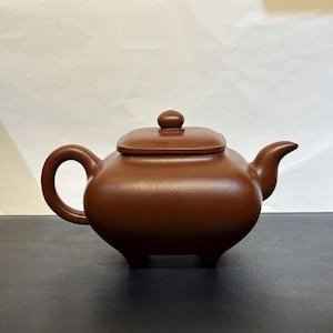 May include: A brown ceramic teapot with a rounded body, a curved handle, and a spout. The lid has a small knob on top. The teapot sits on three small feet. The overall design is simple and elegant.