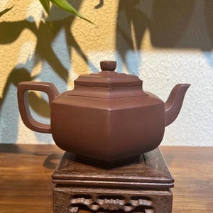 May include: A matte brown clay teapot with a square-shaped body, a flat lid with a small knob, a curved spout, and a rectangular handle. The teapot rests on a dark brown wooden display stand with carved decorative cutouts on the base.