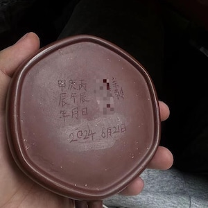 Pode incluir: Uma base de chávena de cerâmica hexagonal castanho-escura com caracteres chineses gravados e a data 2024 6月21日. A superfície tem uma textura mate e terrosa com uma pega visível que se estende da borda inferior do recipiente.