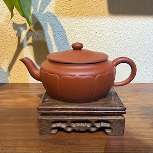 May include: A terracotta-coloured ceramic teapot with a matching lid, placed on a carved wooden stand. The teapot features a curved spout and a rounded handle. The stand is square with decorative cutouts.