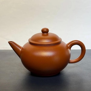 Könnte beinhalten: Eine braune Keramik-Teekanne mit rundem Körper, gebogenem Henkel und Ausguss. Der Deckel hat einen kleinen Knopf oben. Die Teekanne hat eine einfarbige, erdfarbene braune Farbe und scheint aus Ton zu sein.