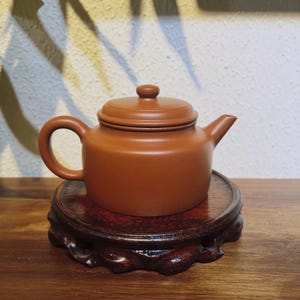 Może przedstawiać: Brązowy ceramiczny czajnik z pokrywką i uchwytem, spoczywający na ciemnej drewnianej podstawce. Czajnik ma zaokrąglony korpus i dzióbek. Podstawka jest okrągła z dekoracyjną, rzeźbioną krawędzią. Tło to neutralna ściana z cieniami.