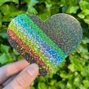Op de afbeelding: Een hartvormige sticker met een glinsterende, holografische afwerking. Het ontwerp heeft een diagonale streep in regenboogkleuren, de rest van het hart is zilverkleurig glitter. De sticker wordt door een hand vastgehouden.