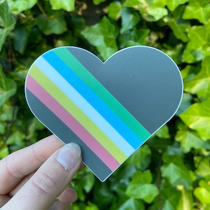 Puede incluir: Una pegatina en forma de corazón con un diseño de rayas diagonales en rosa, amarillo, blanco, azul y verde. La pegatina está sobre un fondo gris oscuro.