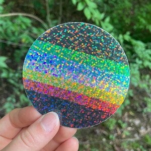May include: A round, iridescent sticker with a rainbow glitter design. The sticker has a black background with five horizontal stripes of glitter in shades of green, blue, purple, red, and orange.