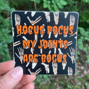 May include: A black sticker with a white skeleton bone pattern. The text "Hocus Pocus My Joints Are Bogus" is printed in orange.