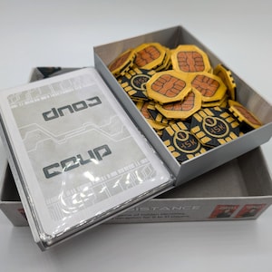 May include: A board game box set with the game title "Coup" visible on the card deck. The set includes a deck of cards and a box filled with yellow and black game tokens. The tokens have a unique design.