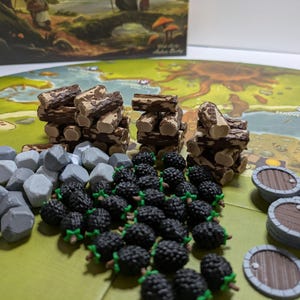 May include: A collection of board game components including three stacks of brown wooden logs, a pile of grey stone tokens, a cluster of black blackberry tokens with green stems, and several round brown wooden barrel tokens on a green game board.