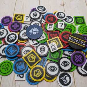 Puede incluir: Una colección de fichas de juego coloridas en varias formas, incluyendo círculos y cuadrados. Las fichas presentan símbolos y la palabra "DECKERS" en una ficha negra y amarilla. Los colores incluyen verde, azul, amarillo, rojo, morado y blanco.