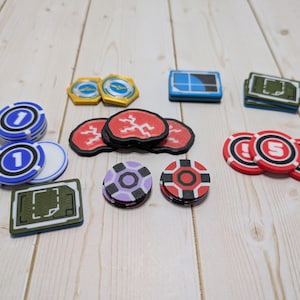 May include: A selection of colourful game chips and tokens, including blue and white chips with the number 1, red and black chips with the number 5, and various other geometric designs in yellow, purple, and green. The items are arranged on a light wood surface.