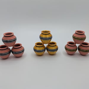 May include: A collection of small, decorative terracotta pots. The pots are painted in shades of pink and yellow, with black and white horizontal stripes. The pots are arranged in groups of three.