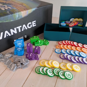 Op de afbeelding: Een bordspelset met een zwarte doos met het label "VANTAGE" en diverse spelstukken. De stukken omvatten blauwe, groene, paarse, grijze, gele, oranje en witte fiches en rechthoekige stukken. Drie teal dozen zijn ook zichtbaar.
