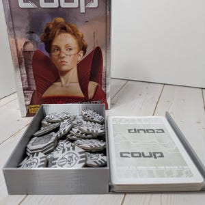 Könnte beinhalten: Das Brettspiel "Coup" wird gezeigt, mit einer Box mit einem Frauenporträt und dem Titel des Spiels. Ein grauer Behälter enthält Spielchips und Karten, ebenfalls mit der Aufschrift "Coup".