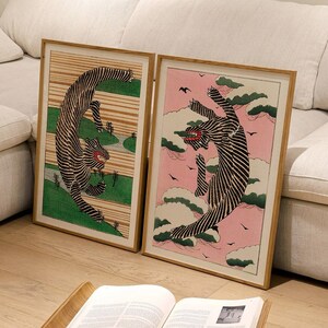 Puede incluir: Dos impresiones enmarcadas de ilustraciones estilizadas de animales en blanco y negro. La impresión de la izquierda tiene un fondo verde y la impresión de la derecha tiene un fondo rosa. Las impresiones están colgadas en una pared blanca sobre un sofá blanco. Un taburete de madera con un libro encima está en primer plano.