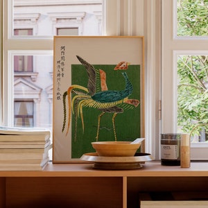 May include: A wooden shelf unit with three shelves, each filled with stacks of magazines and books. A framed print of a colorful bird with Japanese text is on the top shelf, along with a wooden bowl, a candle, and a vase.