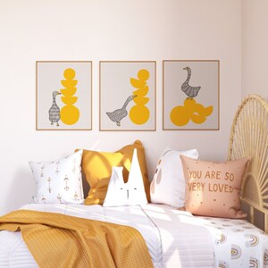 May include: Three framed prints with a minimalist design featuring a gray goose and yellow circles. The prints are hung on a white wall above a bed with yellow and white pillows.