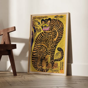 May include: A framed print of a yellow and black tiger with a pink nose and mouth. The tiger is sitting on a gold background with black and white details. The print is framed in a light wood frame.