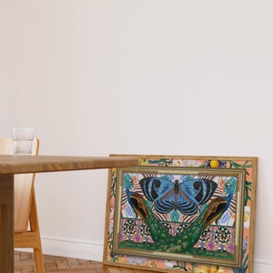 May include: A framed print with a colorful design featuring a large blue butterfly with black markings, two peacocks with green and blue feathers, and a floral border. The background is a peach color with a floral pattern.