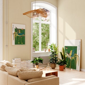 May include: A living room interior with a beige couch, a woven pendant light, and two framed prints of birds on a green background. The prints feature Japanese text. The room has a large window with green trees outside and a few potted plants.