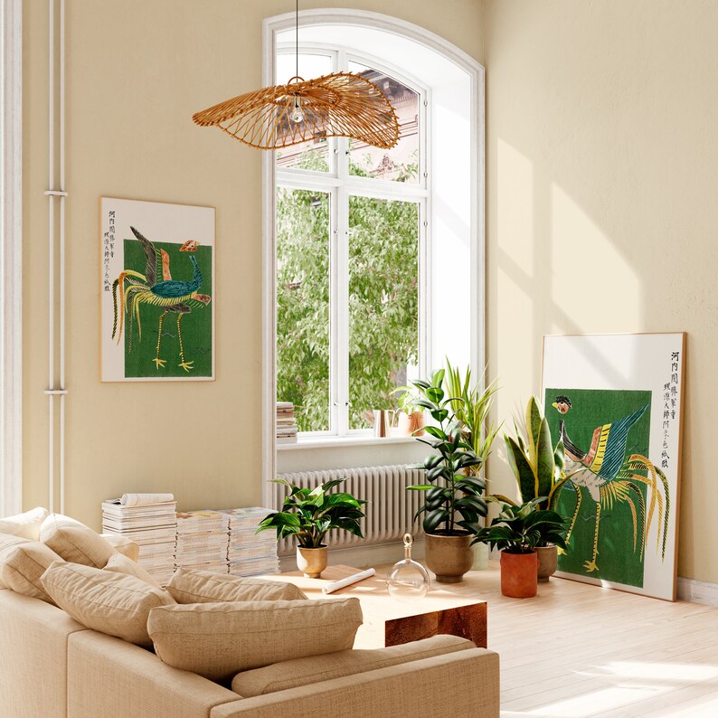May include: A living room interior with a beige couch, a woven pendant light, and two framed prints of birds on a green background. The prints feature Japanese text. The room has a large window with green trees outside and a few potted plants.