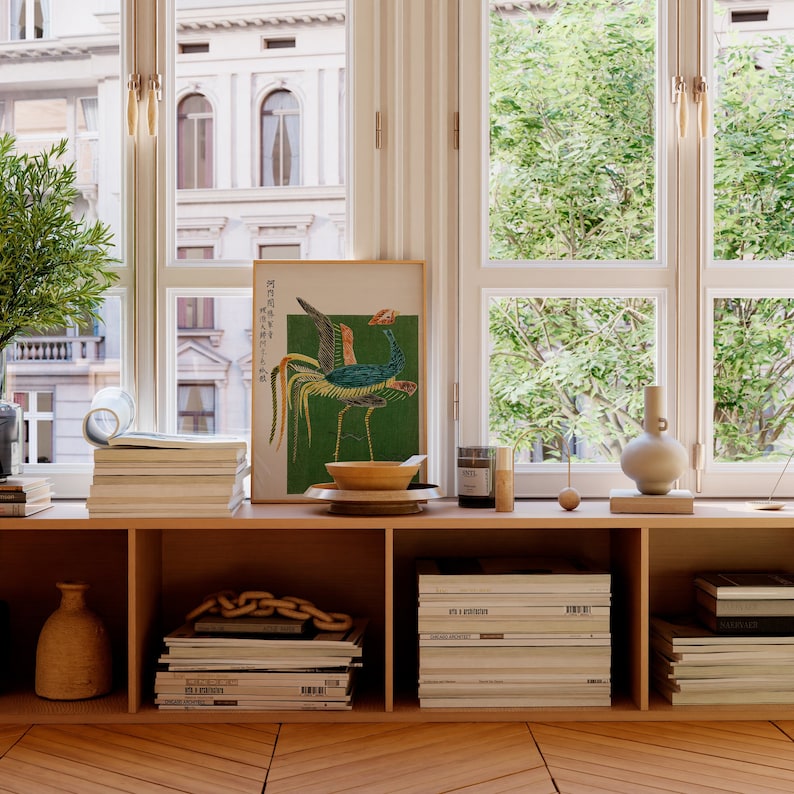 May include: A wooden shelf unit with three shelves, each filled with stacks of magazines and books. A framed print of a colorful bird with Japanese text is on the top shelf, along with a wooden bowl, a candle, and a vase.
