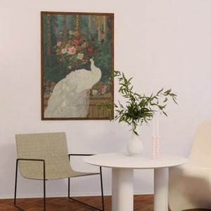 May include: A framed artwork depicts a white peacock with its tail feathers displayed, set against a backdrop of flowers and foliage. A white table with three cylindrical legs, a woven chair, and a vase with greenery and candles complete the scene.