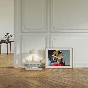 Puede incluir: Una pintura enmarcada de una mujer con un vestido rojo y un sombrero amarillo y azul. La pintura est&aacute; colgada en una pared blanca con moldura blanca. Hay libros apilados en una mesa debajo de la pintura, incluyendo "Live Beautiful" y "Art for Survival".
