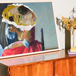 Puede incluir: Una impresi&oacute;n enmarcada de una pintura con un trozo rasgado que revela una pareja bes&aacute;ndose. La pareja est&aacute; representada en un patr&oacute;n amarillo y dorado. La pintura est&aacute; sobre un armario de madera con dos puertas.