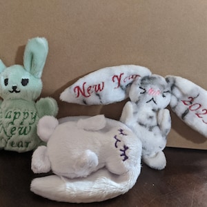 Puede incluir: Tres juguetes de peluche con forma de conejo con detalles bordados. Uno es verde con "Happy New Year" bordado en él. Otro es blanco con marcas negras y grises y "New Year" bordado en una oreja y "2023" en la otra. El tercero es blanco con bordados morados.